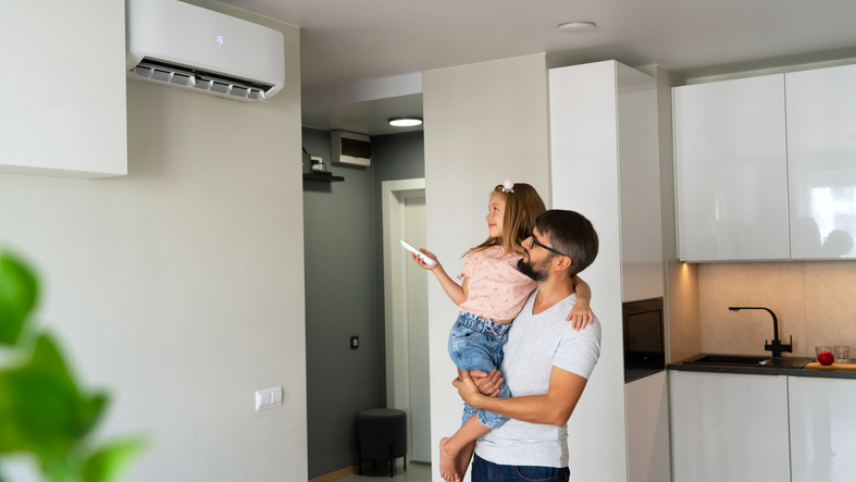 Mann mit Kind auf dem Arm richtet eine Fernbedienung auf ein Split-Klimagerät im Wohnraum