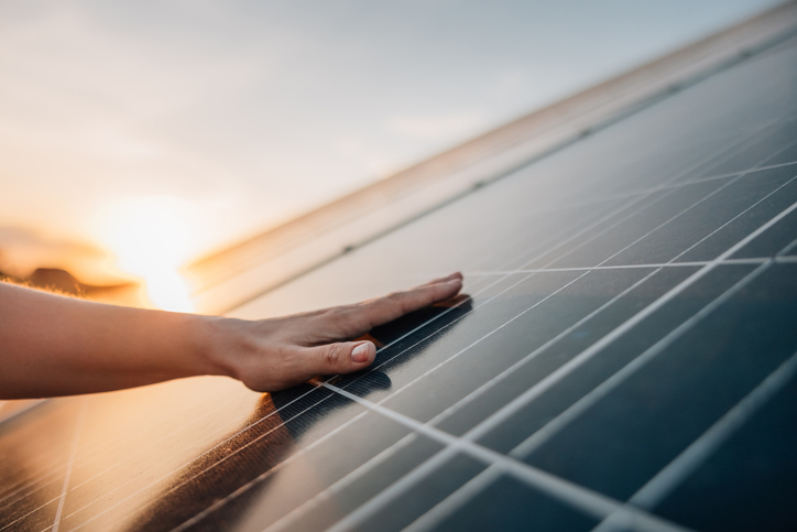 Hand auf einem Photovoltaikmodul, dahinter geht die Sonne unter